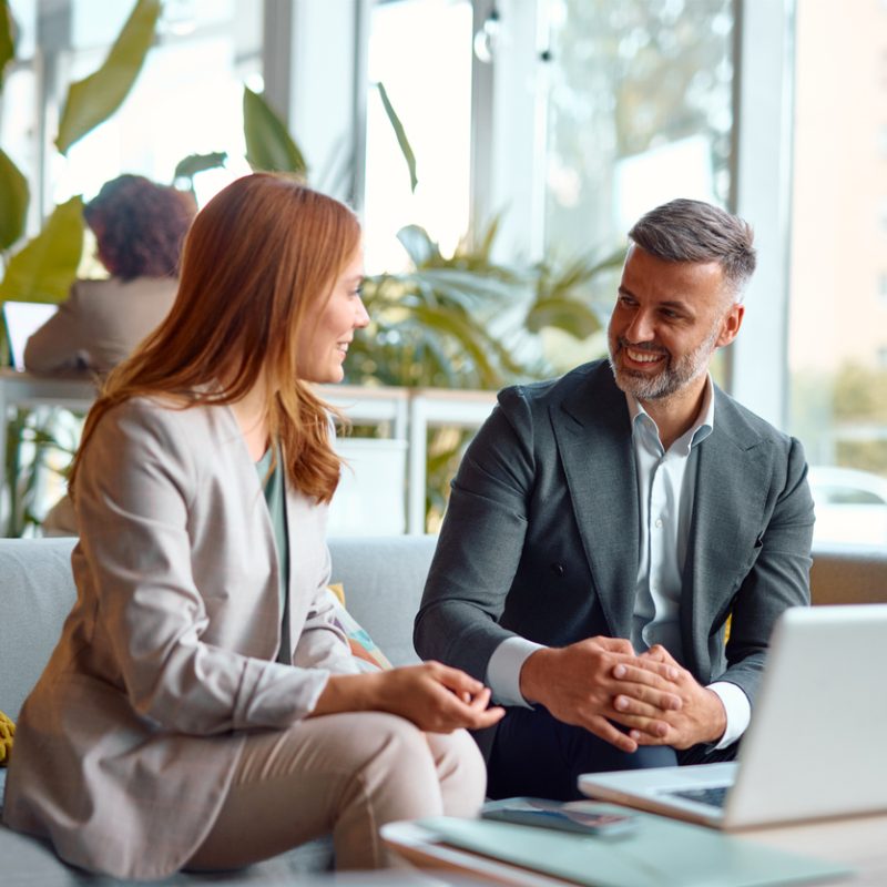 Two business partners are sitting on a couch in a modern office, discussing a project and using a laptop to review data and collaborate on a presentation