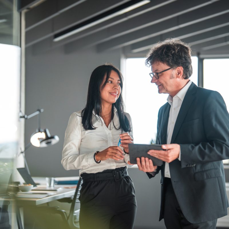 Handsome Hispanic woman working with a mature male coworker in a financial office. They are wearing formal businesswear and using a digital tablet.