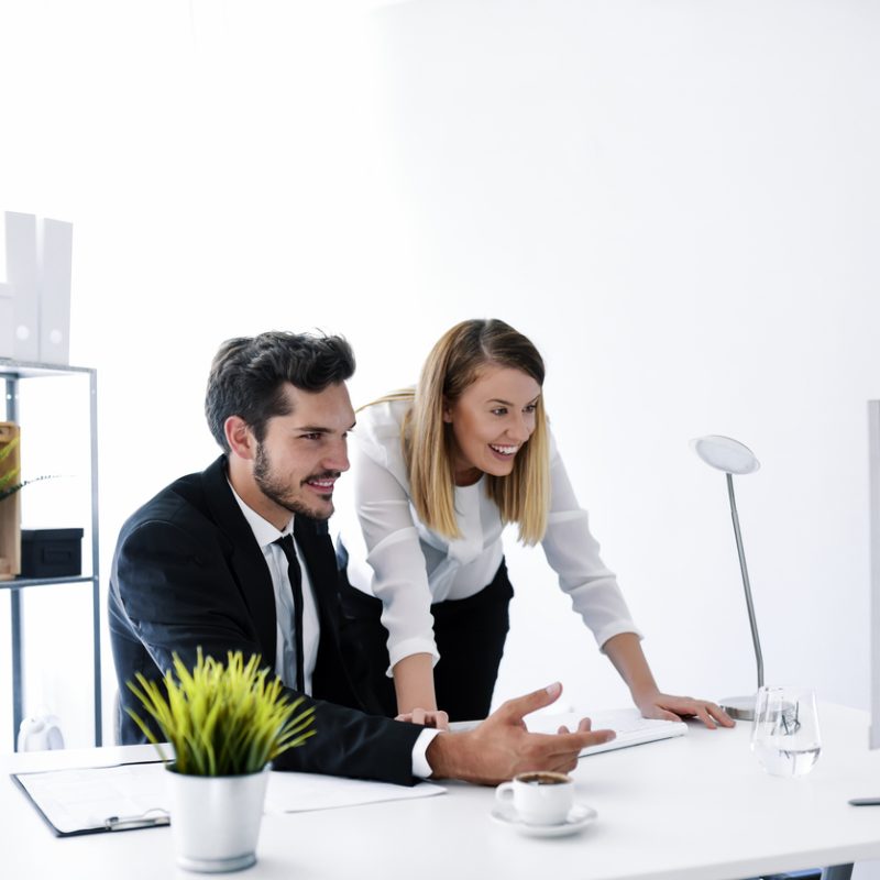 Business, startup and people concept - happy Caucasian creative team with PC computer in white office working together. Focusing on work. Handsome young man and brown hair female looking on monitor and typing something while sitting on working place in creative office.