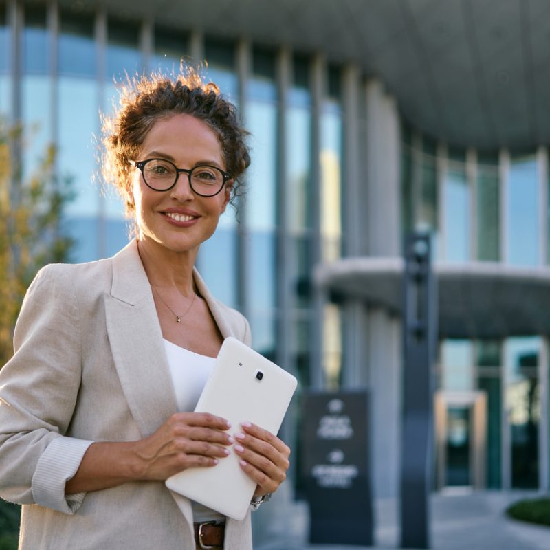 Confident businesswoman with curly hair and eyeglasses holding a digital tablet. Smiling at the camera while standing in front of a contemporary corporate building. Representing success and technology